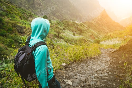 Alone traveler with backpack walking down cobbled trekking trail towards green valley. Sun flares on