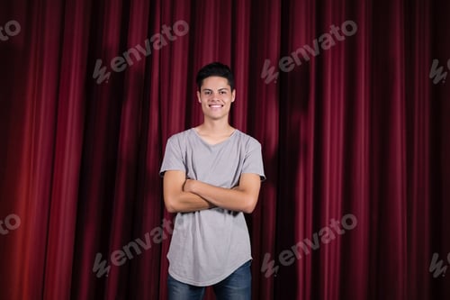 Preview: Hispanic man standing with arms crossed on stage wearing tee and jeans before maroon-red curtains