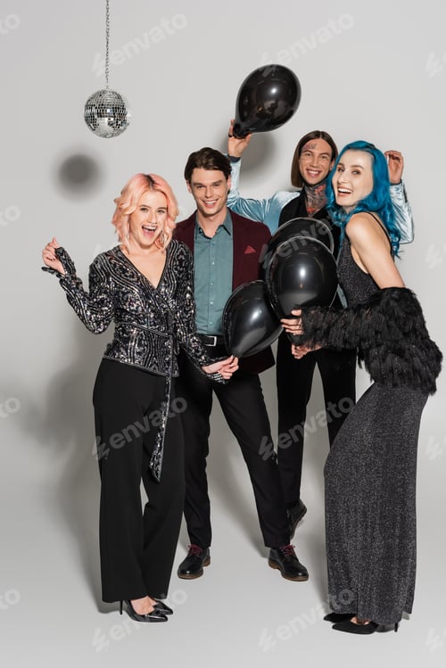 Preview: full length of cheerful and stylish nonbinary friends with black balloons looking at camera on grey