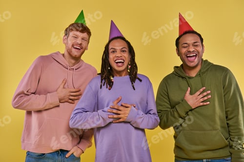 Preview: young multiethnic friends in casual outfits and birthday hats laughing sincerely, yellow backdrop
