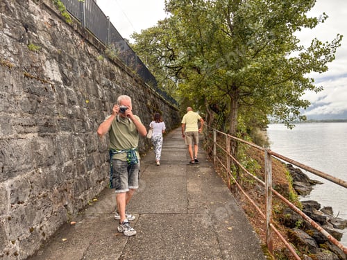 Preview: elderly man walking along Lake Geneva with friends looks back and takes a photograph with his camera