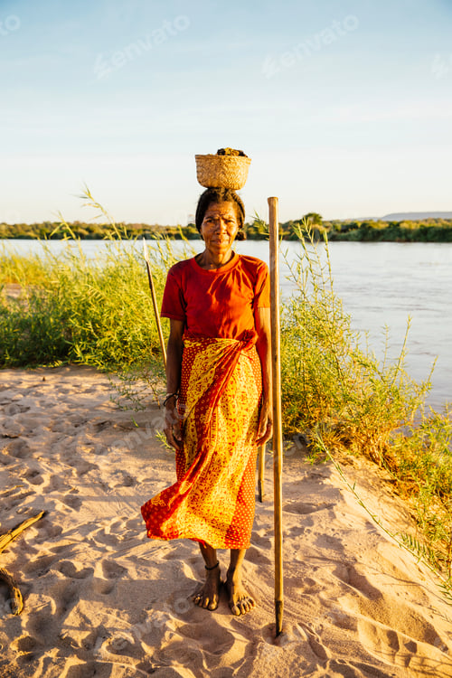 Preview: A woman stands by the riverbank at sunset, balancing a basket on her head.