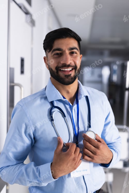 Preview: Portrait of happy biracial male doctor with stethoscope in hospital corridor