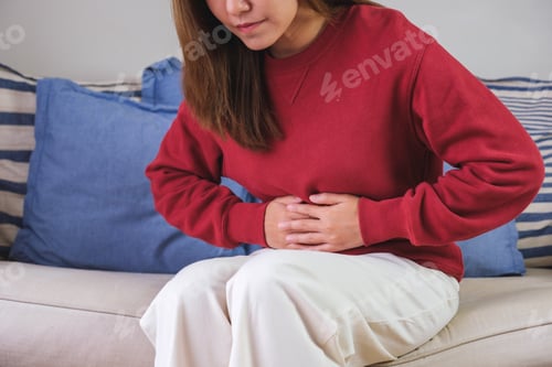 Preview: A sick woman suffering from stomachache, abdominal pain while sitting on sofa at home