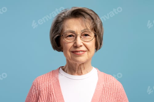 Preview: Portrait of smiling senior woman wearing glasses and pink sweater on blue background