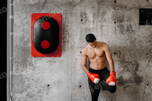 Preview: Brutal dark-haired guy with a naked torso and the red boxing gloves stands against a concrete wall