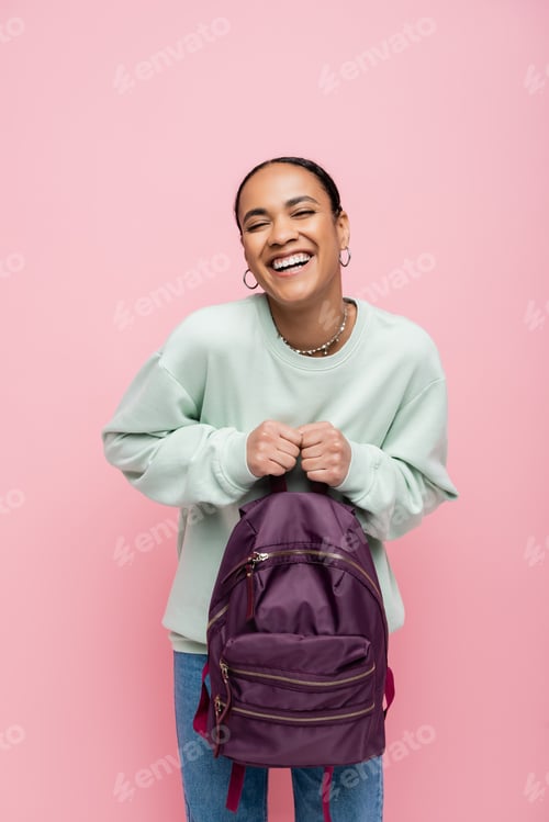 Preview: joyful african american student in sweatshirt holding purple backpack isolated on pink