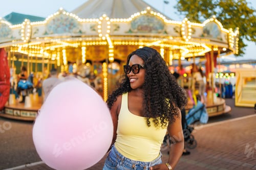 Preview: Young black woman smiling, holding pink cotton candy at a glowing carousel