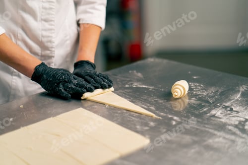 Preview: Close-up shot of a chef's hand in gloves rolling raw cut dough into croissant shapes for baking