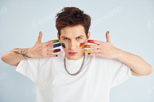 Preview: Young gay man is standing in the studio and posing for a camera. Painted by multi colored makeup