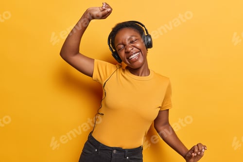 Preview: Horizontal shot of cheerful dark skinned woman dances carefree shakes arms feels positive wears