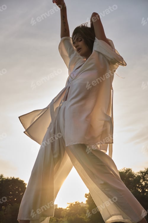 Preview: A woman in elegant white attire dances with her arms lifted, bathed in the warm glow of sunset. The