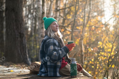 Preview: Contented woman enjoying hot tea from thermos in warm clothes sitting on eco path in autumn forest