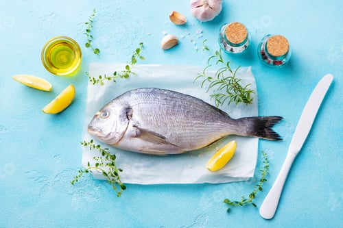 Preview: Dorado Fish with Cooking Ingredients on a Parchment Paper. Blue Stone Background. Top view.