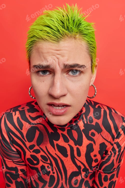 Preview: Puzzled unusual female teenager with trendy green hairstyle looks unhappily at camera frowns face be