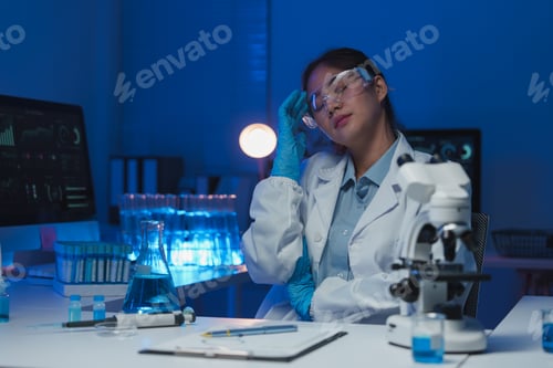 Preview: Young overworked asian female scientist wearing lab coat and gloves is touching her head and sufferi