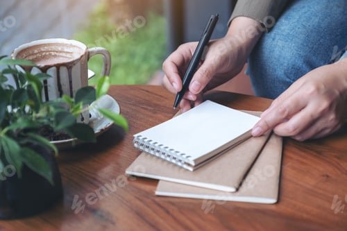 Preview: Closeup image of a woman's hand preparing to write on blank notebook with coffee cup on wooden table