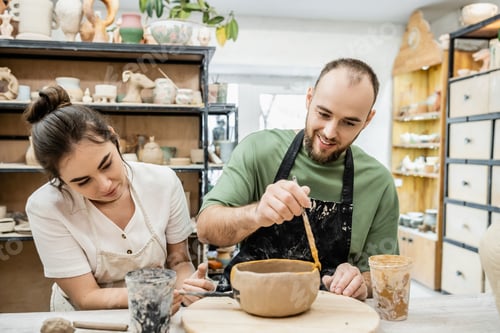 Preview: Smiling craftsman coloring clay bowl near girlfriend in ceramic workshop at background