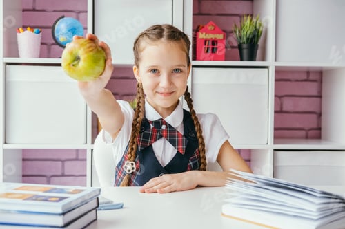 Preview: Little girl a school student sits at her desk and holds out an apple