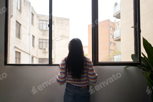 Preview: Rear view African girl leaning out of the window, looking outside where there are large buildings.