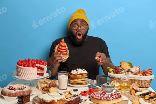 Preview: Stupefied black man eats tasty croissant, points at table full of sweet delicious desserts, wears ha