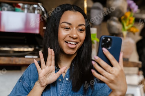 Preview: Black smiling woman gesturing while making video call in cafe
