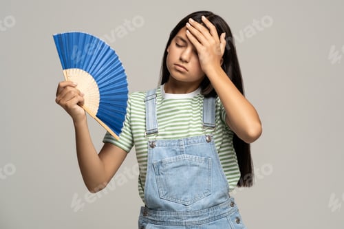 Preview: Sweaty teenage girl touching forehead using paper fan suffer from heat cooling in hot summer weather