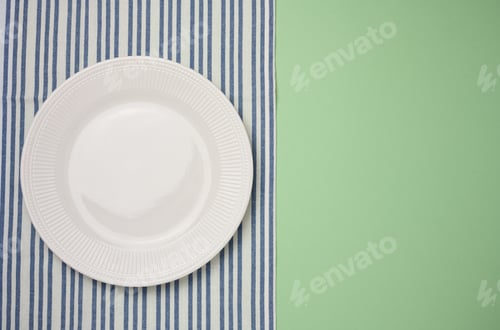 Preview: Empty round ceramic plate and blue kitchen towel on green background, top view