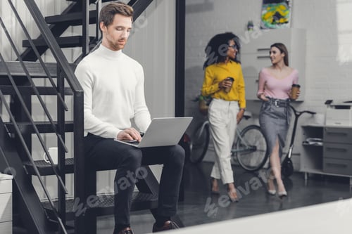 Aperçu: focused young man using laptop on stairs and businesswomen walking behind in office