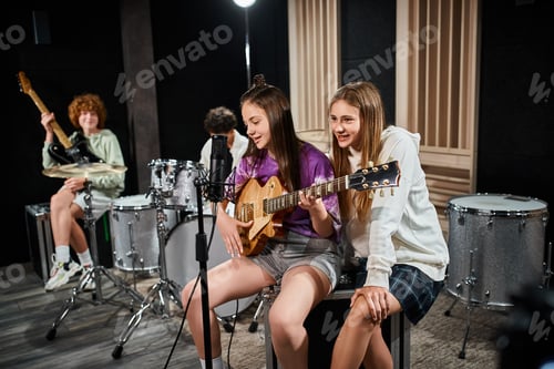 Preview: focus on teenage girls with guitar singing with blurred friends with drums and guitar on backdrop