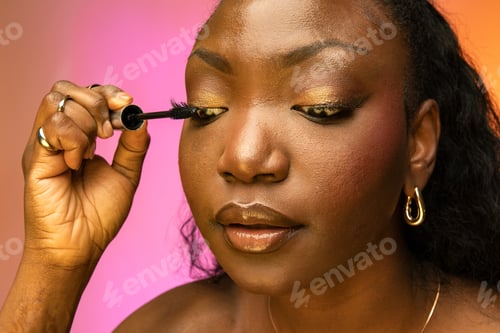 Preview: Beautiful African American woman doing makeup with cosmetic mascara brush in hand in studio
