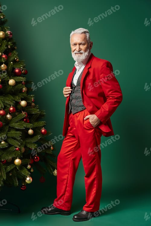 Preview: classy Santa in red suit with white beard posing near fir tree with hand in pocket, winter concept