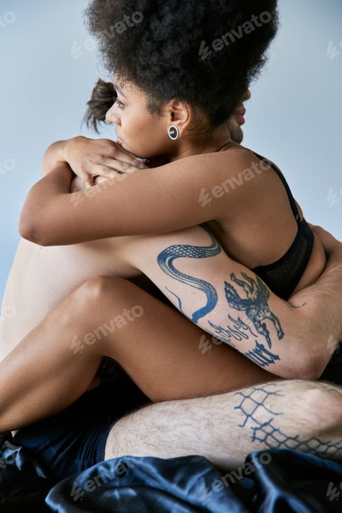 Preview: beautiful young multicultural couple sitting on their bed and hugging lovingly on gray backdrop