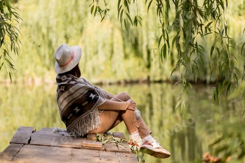 Preview: young beautiful girl in fashionable clothes enjoys nature. Sits on the bridge by the lake