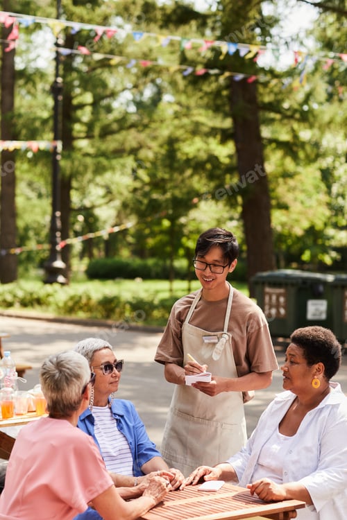 Preview: Vertical smiling young waiter serving group of senior women at outdoor cafe
