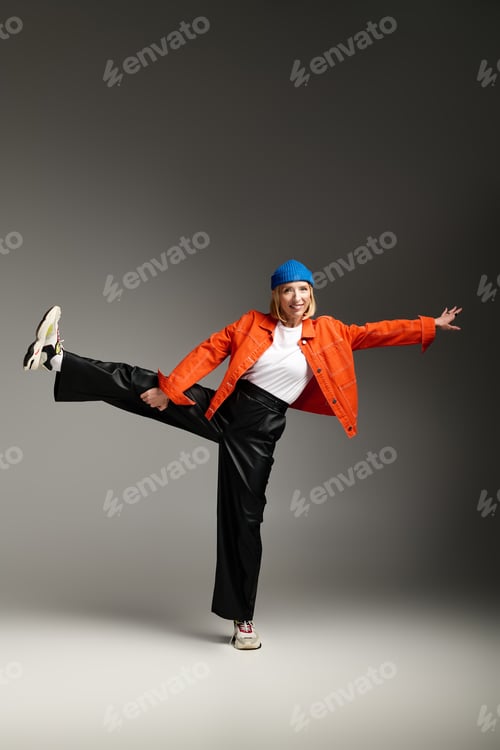 Preview: Charming blonde woman in a blue beanie and orange jacket poses stylishly in a studio