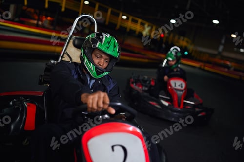 Preview: african american driver in helmet driving go kart on circuit near friend on blurred backdrop