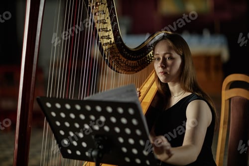 Preview: Portrait of Female Harpist Turning Page with Notes Performing Music in Dim Light Indoors