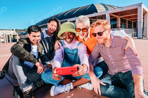 Preview: Multiethnic group of friends taking pictures together sitting outdoors under blue sky.