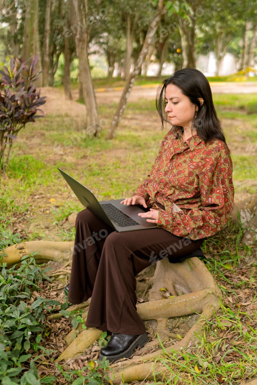 Preview: Young woman remotely working on laptop in park