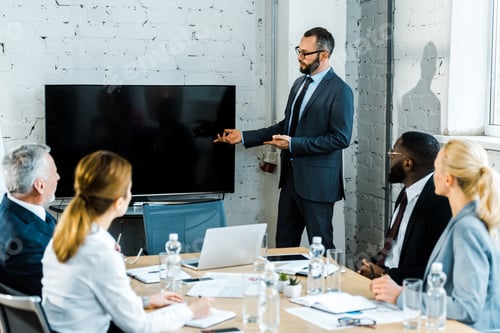 Preview: bearded business coach in eye glasses gesturing near tv with blank screen and multicultural