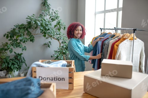 Preview: Young smiling girl choosing and folding clothes in charity boxes.