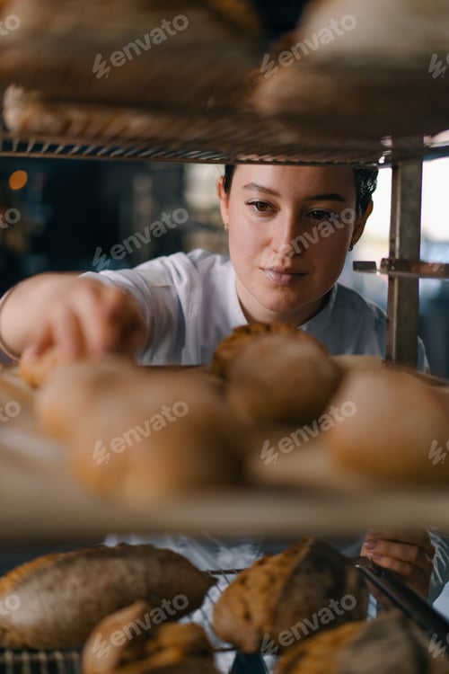Preview: attractive female baker between shelves looking and checking freshly baked bread very carefully