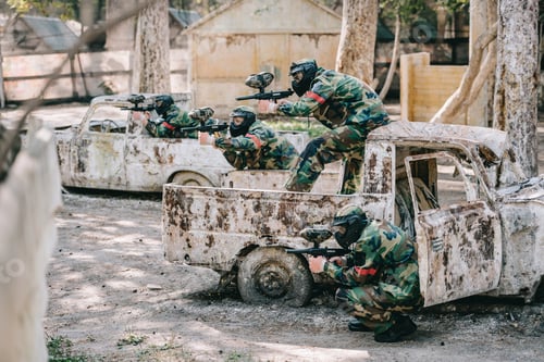 Preview: paintball team in camouflage and goggle masks aiming with marker guns from broken cars outdoors
