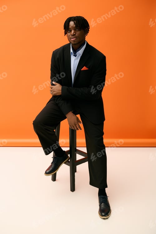 Preview: Handsome young man poses confidently in stylish attire against a vibrant orange backdrop