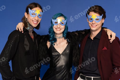 Preview: happy queer person in black leather dress embracing nonbinary friends in party masks isolated on