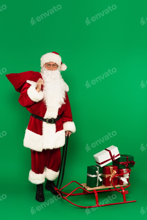 Preview: Santa claus with sack standing near sleigh with gifts on green background