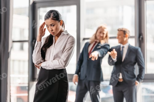 Preview: frustrated asian businesswoman standing in office, businessmen behind gesturing and laughing