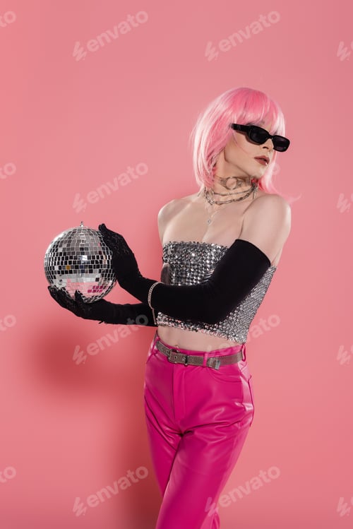 Preview: Fashionable drag queen in sunglasses and silver top holding disco ball on pink background