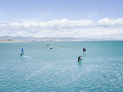 Preview: Panoramic photo of several people windsurfing in the sea with the mountains in the background.